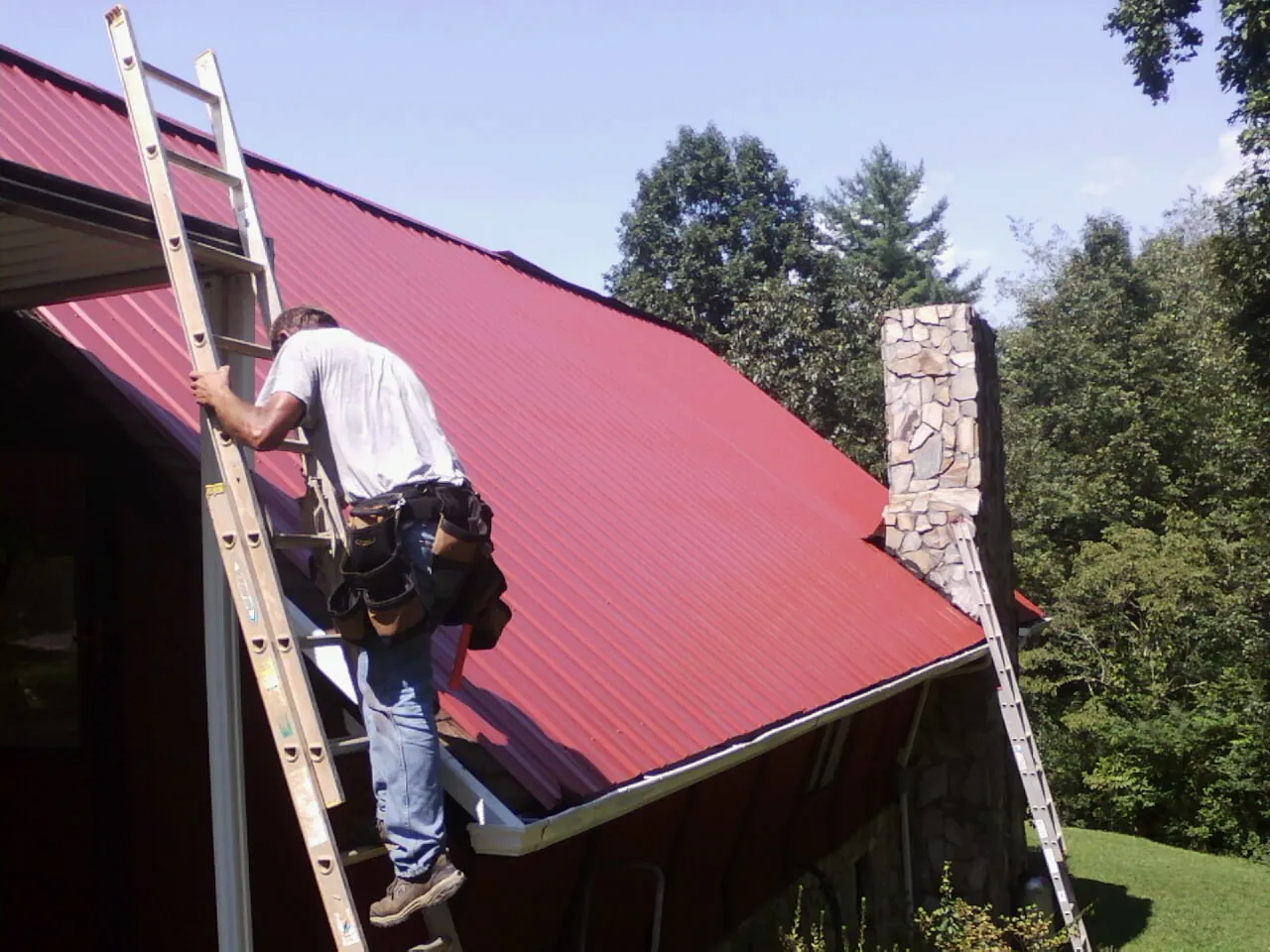 Roofer on a ladder beside a red metal roof and stone chimney — Soffit & Fascia Repair by Roofing Brothers in Fond Du Lac